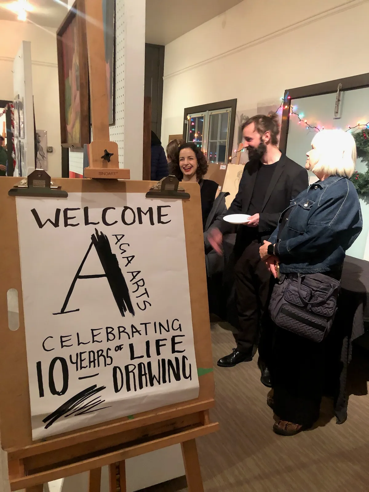 A hand-lettered welcome sign on an easel reads "Welcome, AGA Arts, celebrating 10 years of life drawing" as guests arrive.