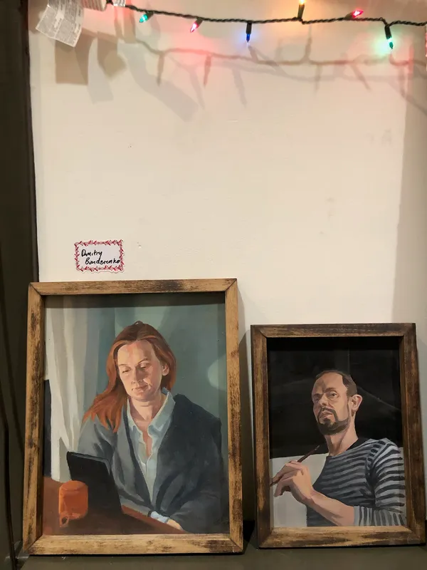 Two framed oil portraits by Dmitry Bondarenko propped against a wall under string lights: a woman reading, and a man holding a paintbrush.