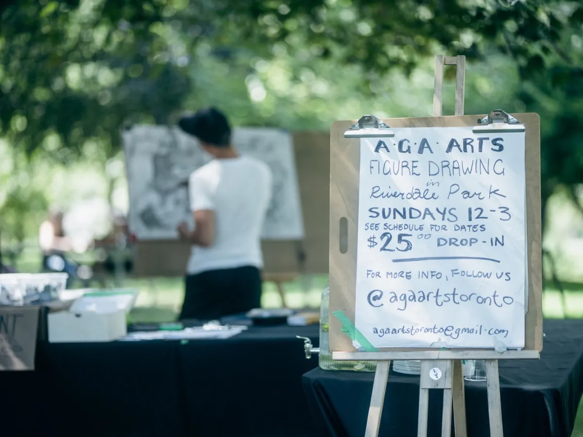 An easel sign reads "AGA Arts figure drawing in Riverdale Park, Sundays 12–3, $25 drop-in, @agaartstoronto" in front of an artist at work.