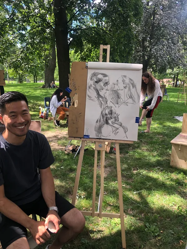 A smiling artist sits beside his easel, which holds charcoal portraits of the model.
