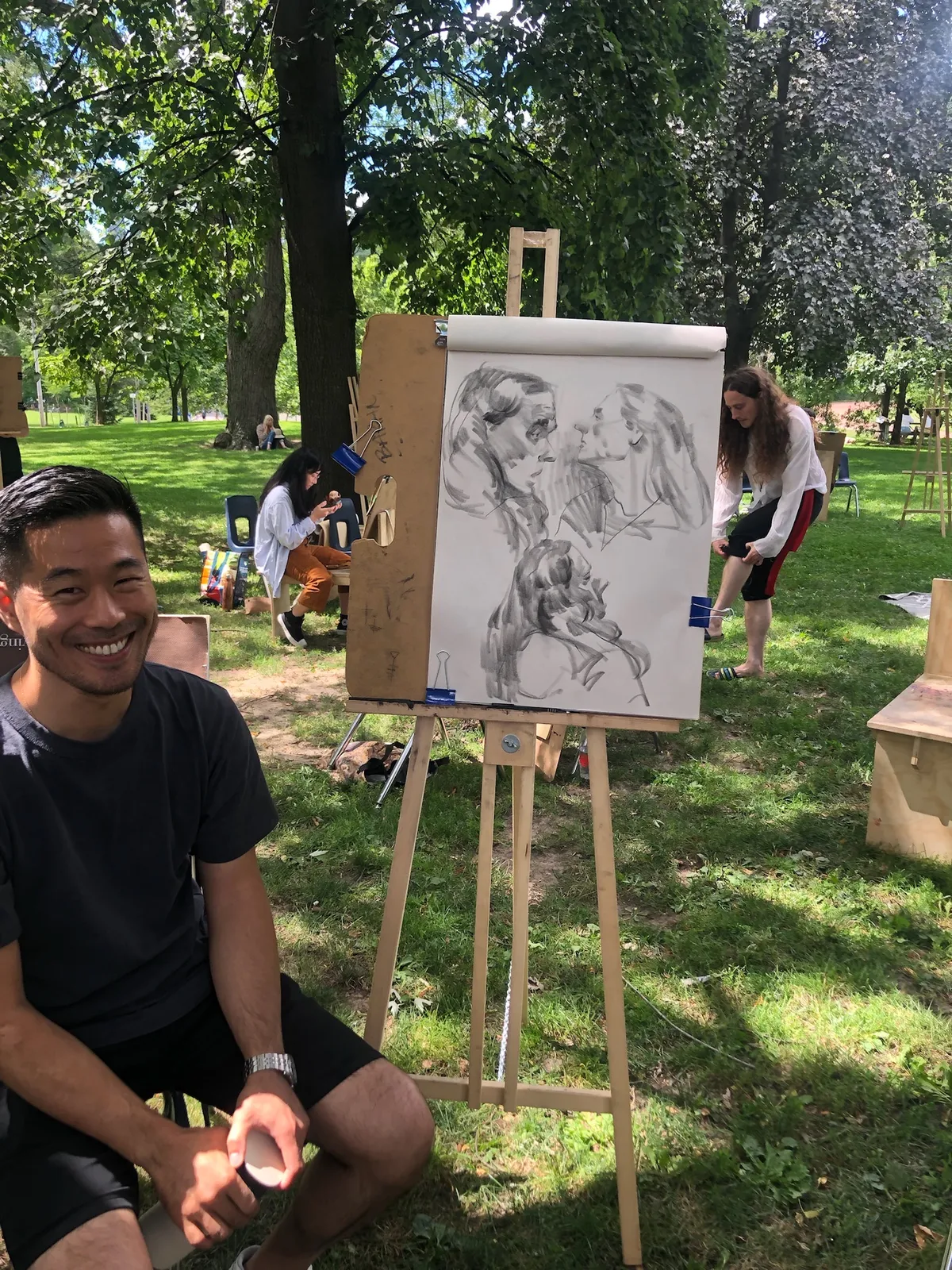 A smiling artist sits beside his easel, which holds charcoal portraits of the model.