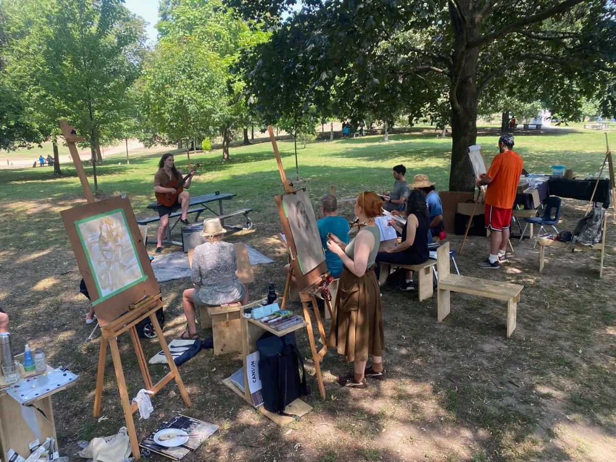 Artists paint at easels in the shade while a guitarist plays at a picnic table.