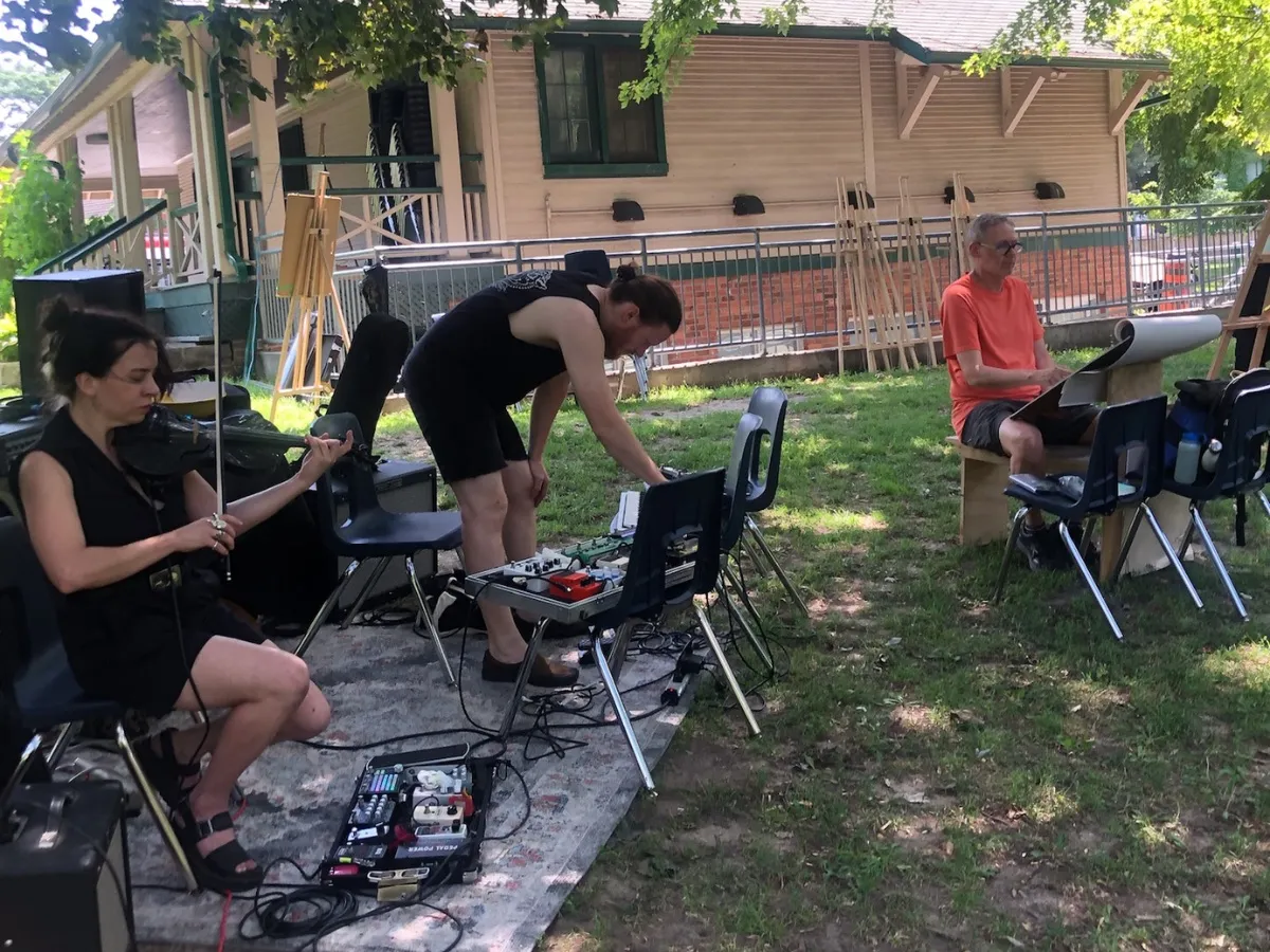 Two musicians play on a rug in the park, one on violin and one on a row of effects pedals.