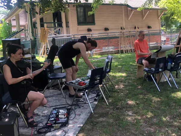 Two musicians play on a rug in the park, one on violin and one on a row of effects pedals.