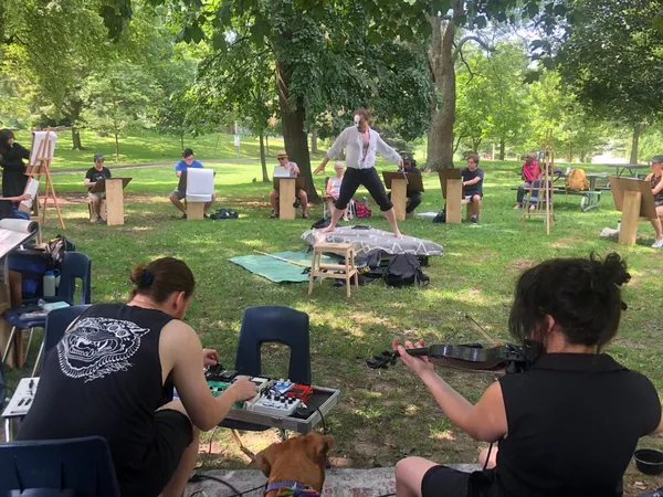 A model in a white shirt poses with arms outstretched, surrounded by artists, as musicians play in the foreground and a dog looks on.