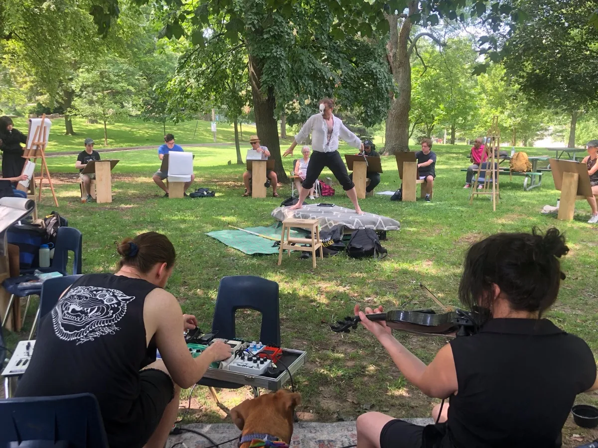 A model in a white shirt poses with arms outstretched, surrounded by artists, as musicians play in the foreground and a dog looks on.