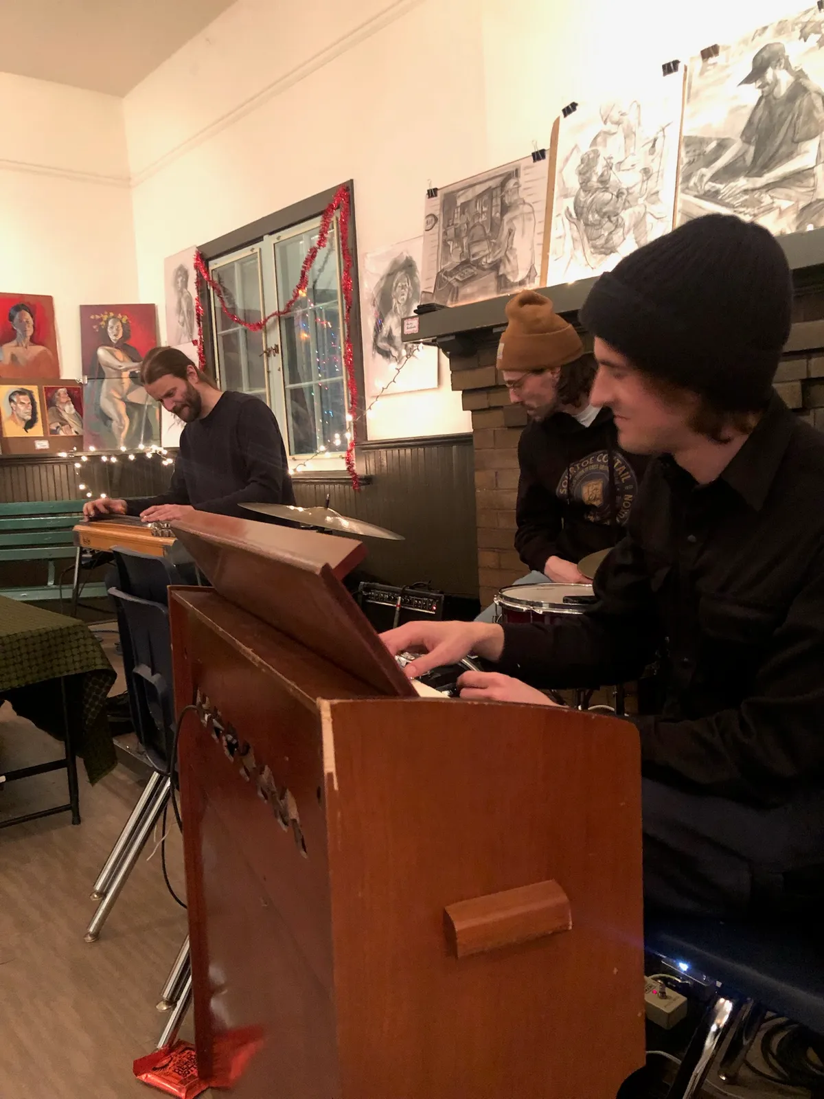 Three musicians play keyboard, drums and a small organ in a room hung with figure drawings and paintings.