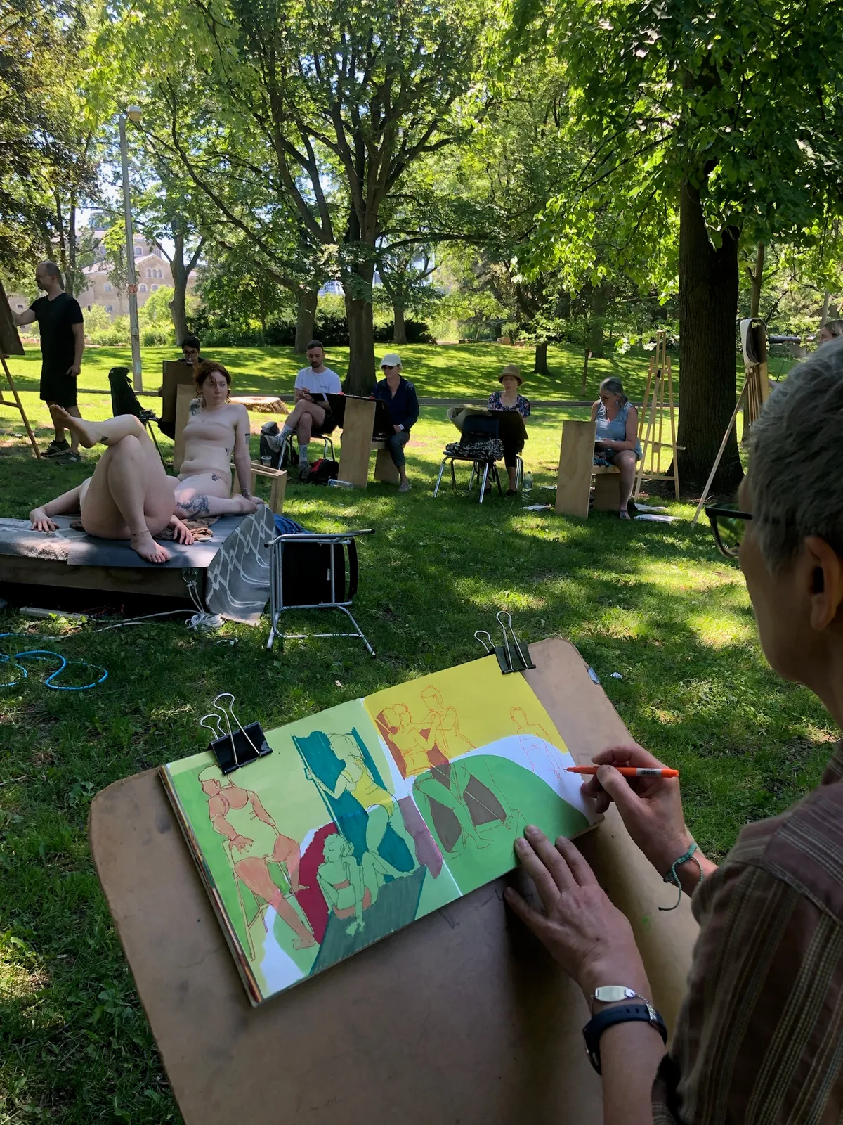 An artist in the foreground colours a sketchbook page of figures while models pose on a platform in the park.