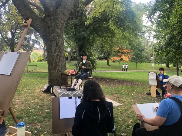 A model in 18th-century costume and tricorn hat sits beside a table of flowers as artists draw.