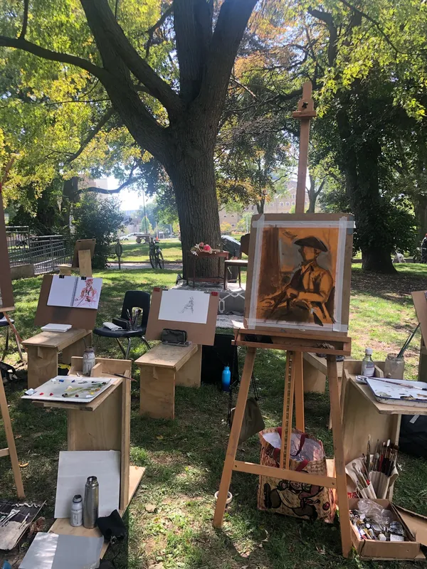 Easels and drawing benches set up under a large tree, one holding a chalk portrait of the costumed model.