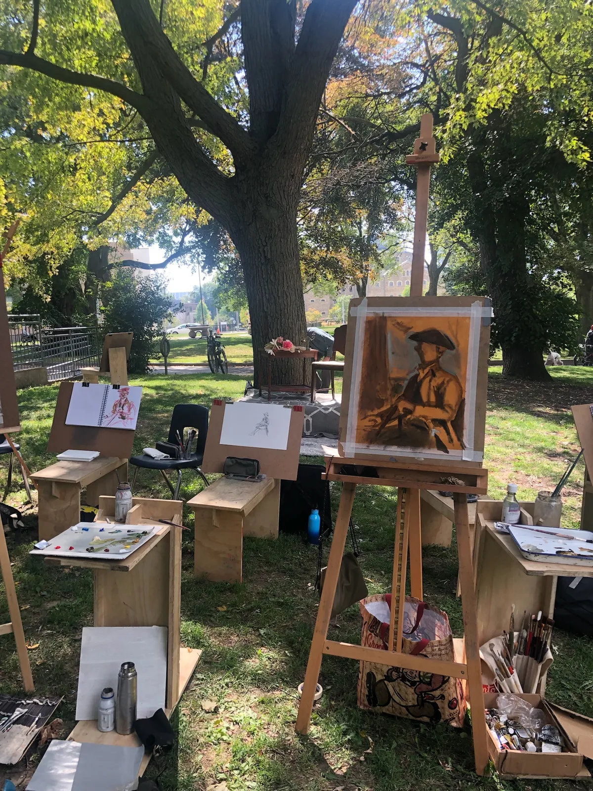 Easels and drawing benches set up under a large tree, one holding a chalk portrait of the costumed model.