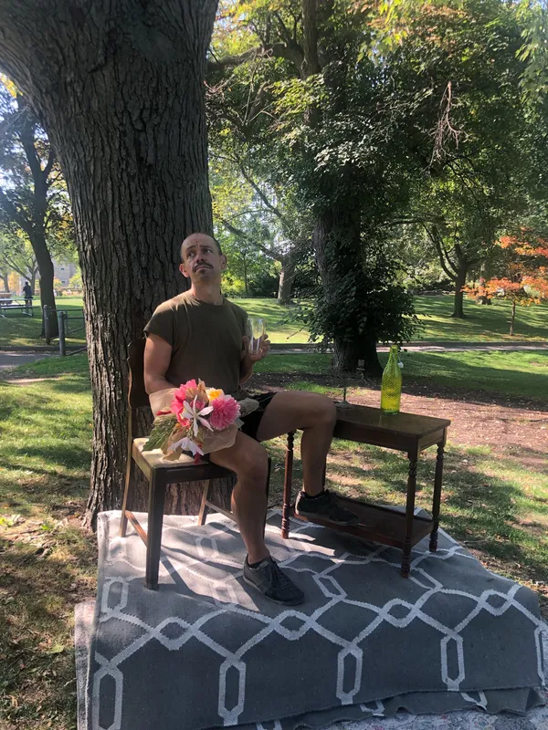 A model sits on a wooden chair under a tree, holding a bouquet of flowers, beside a small table with a bottle.