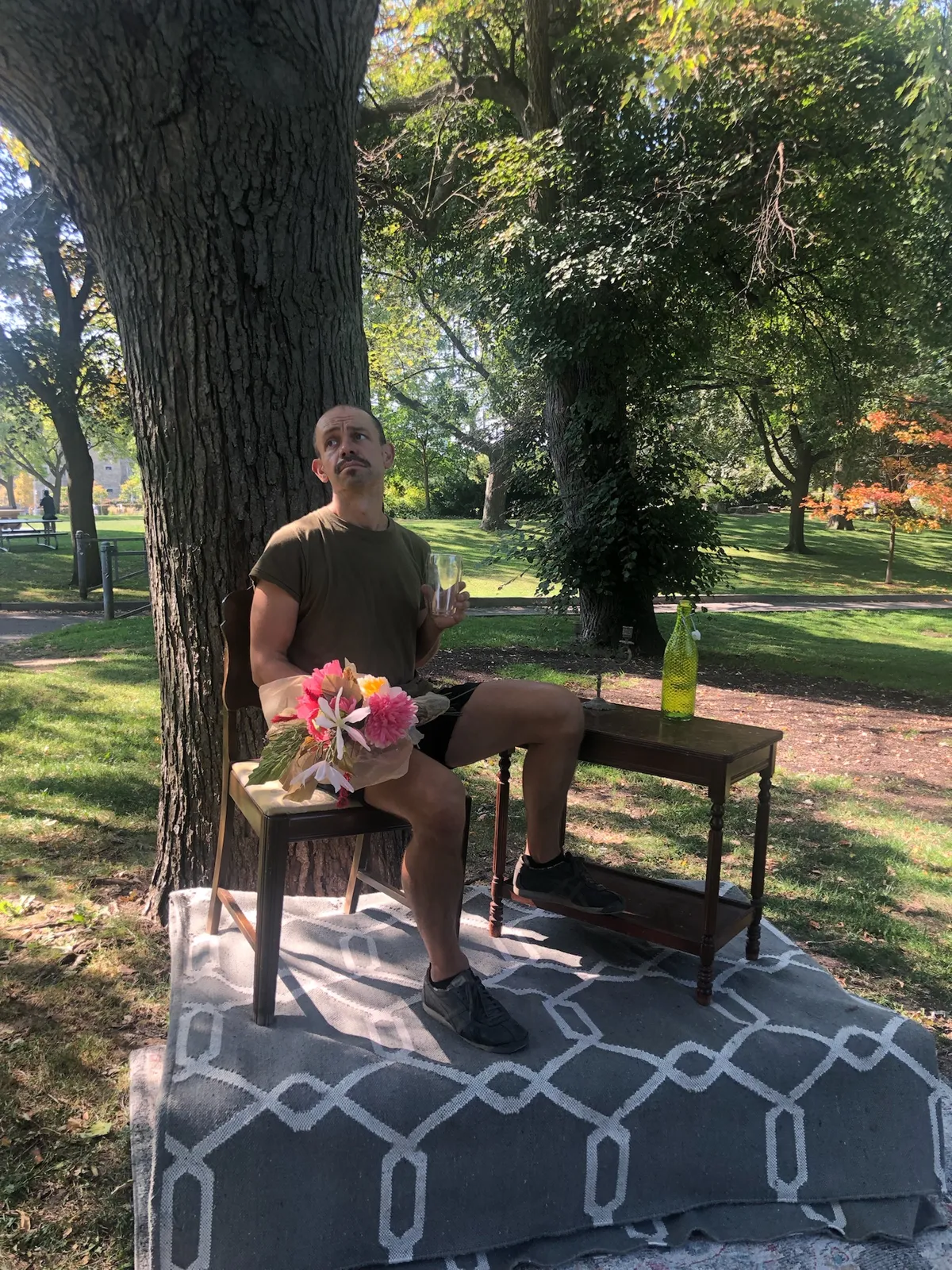 A model sits on a wooden chair under a tree, holding a bouquet of flowers, beside a small table with a bottle.