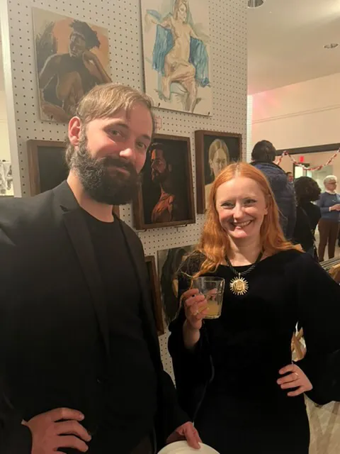 Two guests pose smiling in front of a pegboard wall of framed figure paintings.
