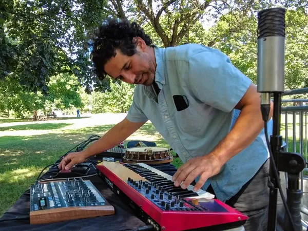 A musician plays a red keyboard and a synthesizer under the trees, beside a microphone.