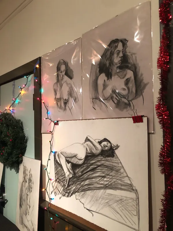 Charcoal nude studies pinned to a wall strung with Christmas lights and tinsel.