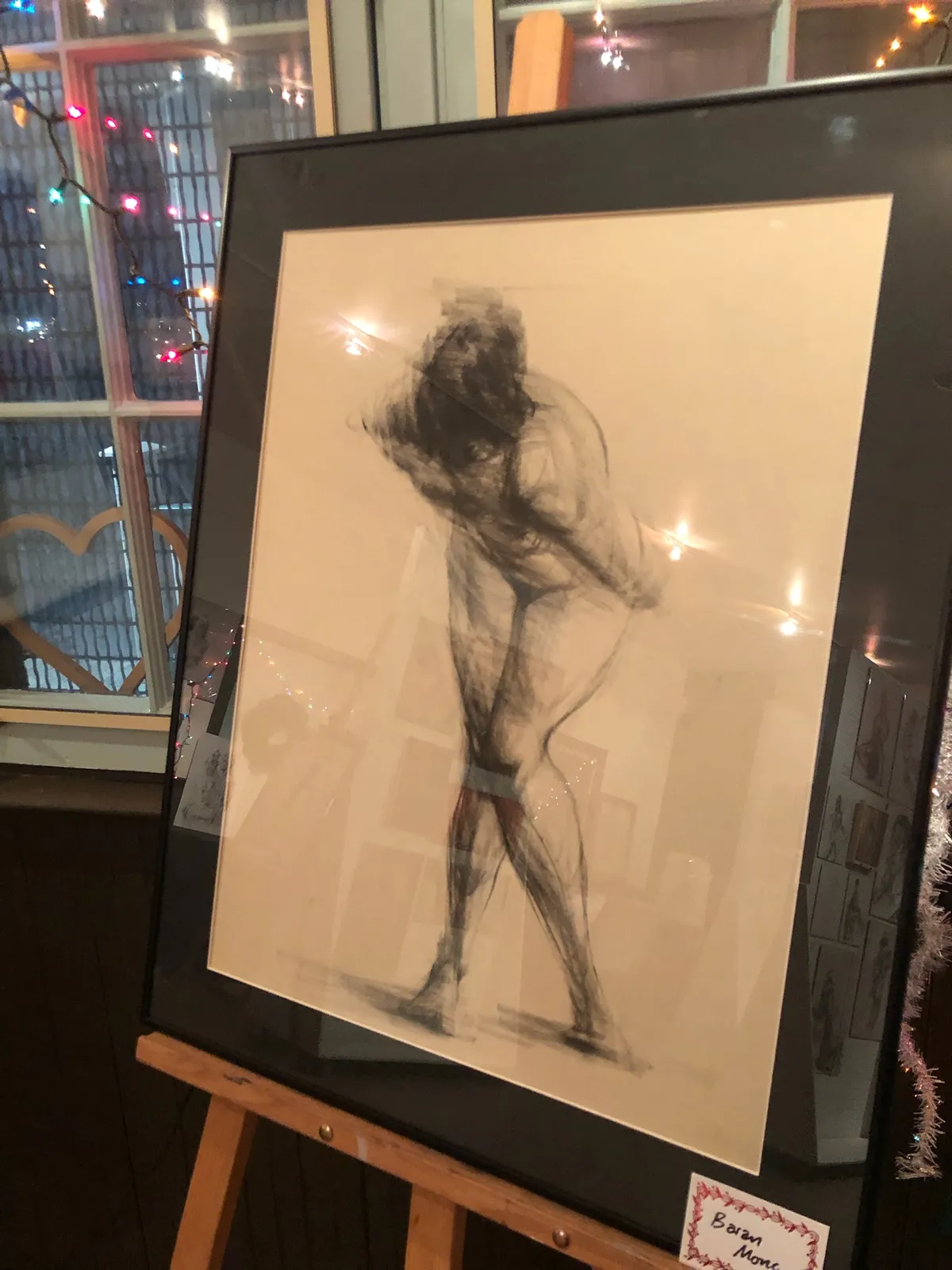 A large framed charcoal drawing of a standing nude figure, on an easel by a window.