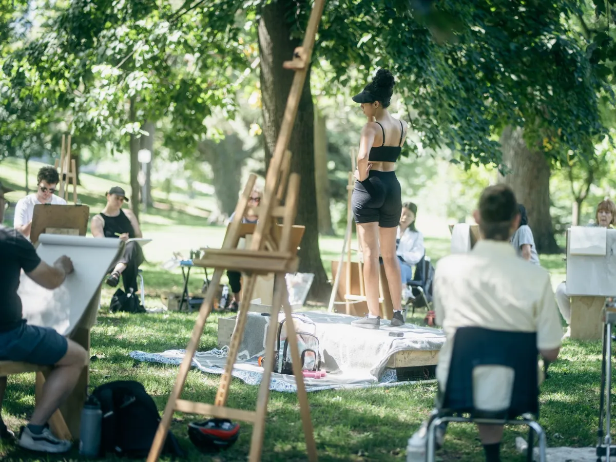 A costumed model stands on a platform under a tree, surrounded by artists at easels and drawing horses.