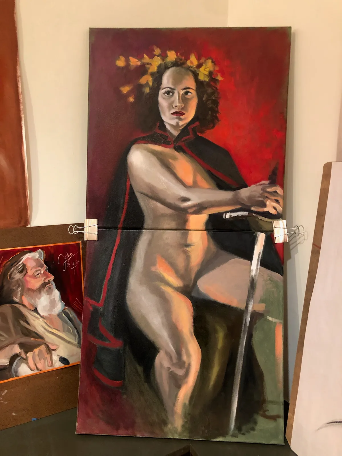 A large oil painting of a nude woman with a laurel wreath, cloak and sword, beside a smaller portrait of a bearded man.
