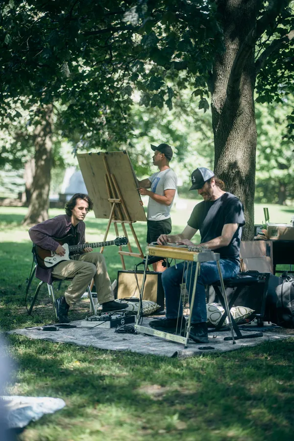 Two musicians, one on guitar and one on pedal steel, play beside an artist at an easel.