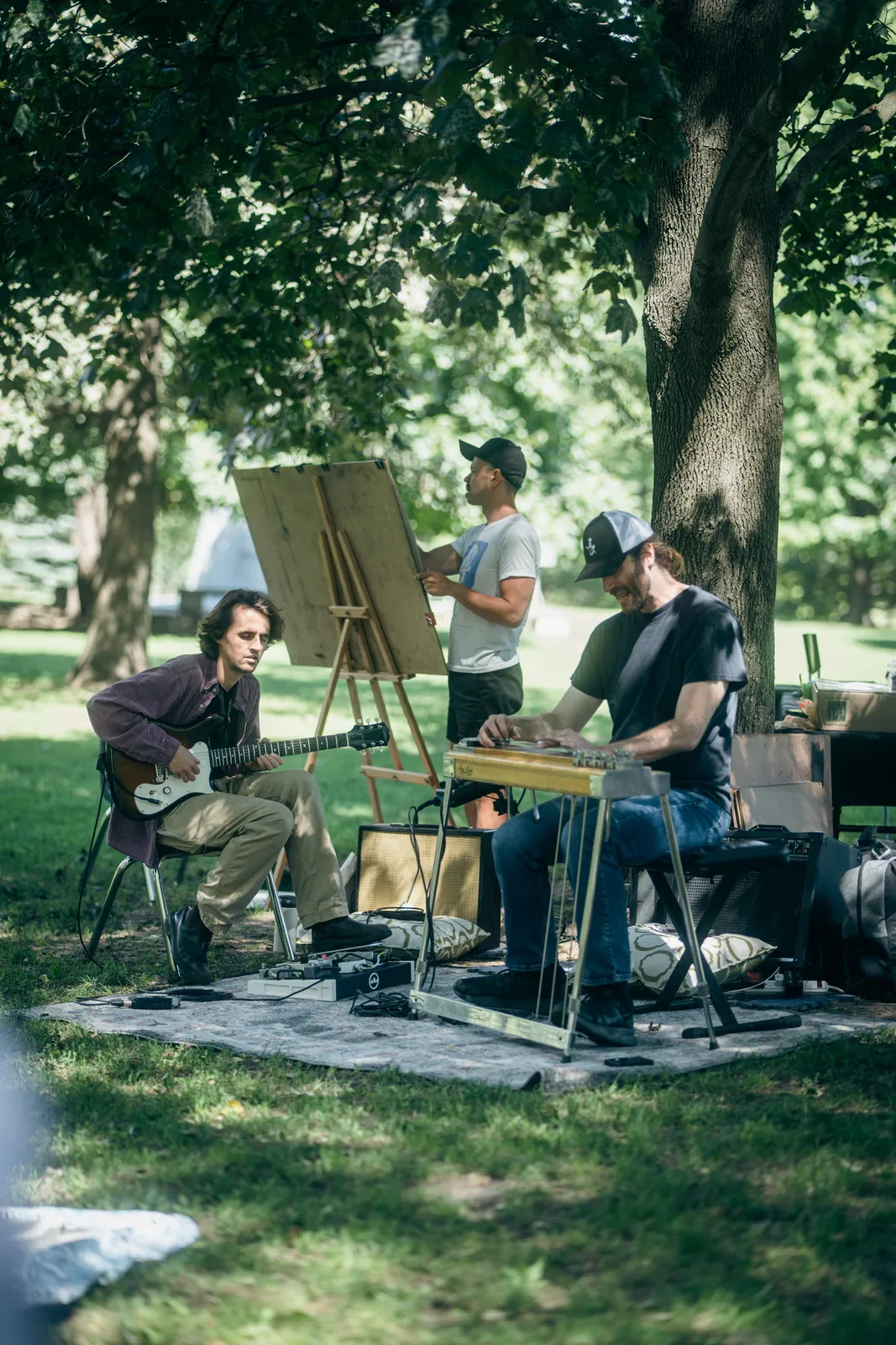 Two musicians, one on guitar and one on pedal steel, play beside an artist at an easel.